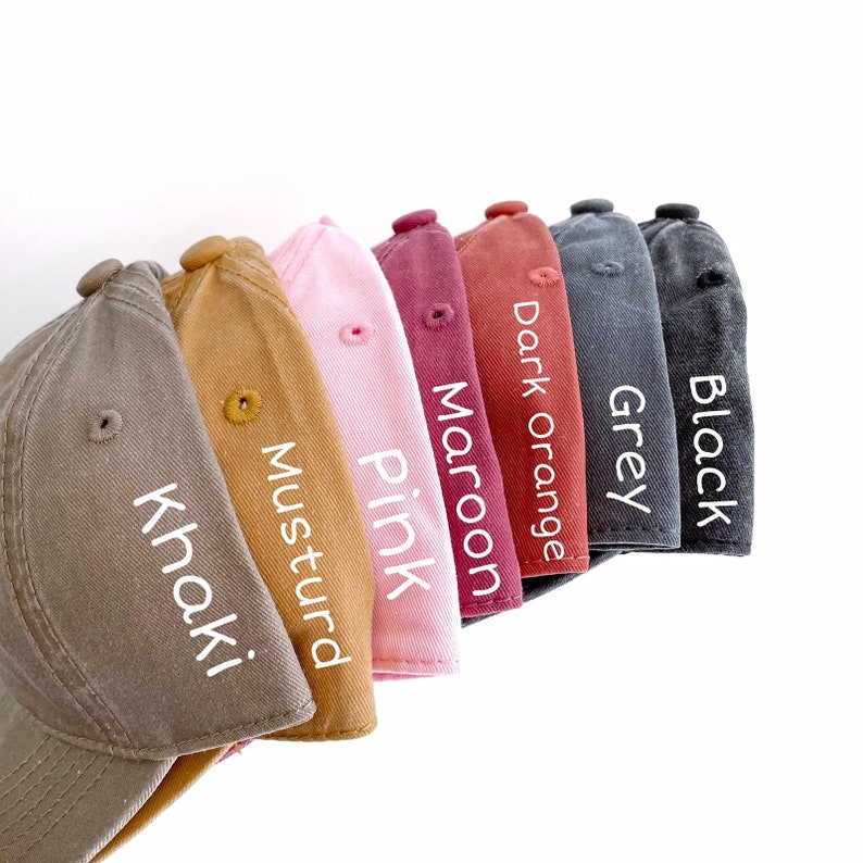 Puede incluir: Una fila de nueve gorras de b&eacute;isbol de diferentes colores, incluyendo caqui, mostaza, rosa, granate, naranja oscuro, gris y negro. Cada gorra tiene un color diferente y est&aacute; etiquetada con su nombre de color.