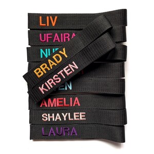 Op de afbeelding: Een stapel zwarte stoffen banden met geborduurde namen in diverse kleuren, waaronder oranje, roze, turquoise, geel, lichtroze, lichtblauw, rood, zachtroze en paars. De namen LIV, UFAIRA, NURI, BRADY, KIRSTEN, AMELIA, SHAYLEE en LAURA zijn zichtbaar.