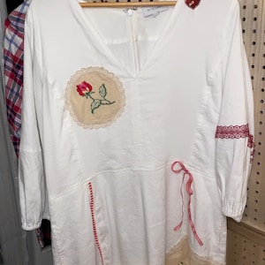 May include: White dress with a V-neck and three-quarter sleeves. Features embroidered floral patches, red trim, and a heart-shaped applique. The dress is hanging on a wooden hanger.