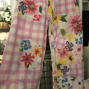 May include: Pink and white gingham trousers with floral patches. The trousers have two large pockets on the front and two smaller pockets on the back.