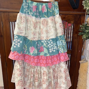 Puede incluir: Una falda larga y de capas con estampado floral. La falda está hecha de diferentes telas y tiene un diseño de patchwork. La falda es en tonos de verde, rosa y crema.