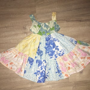 May include: A sleeveless dress featuring a patchwork design of various floral fabrics in shades of blue, yellow, pink, and green. The bodice has a scoop neckline and the skirt is constructed with multiple panels of patterned cotton-like fabric.
