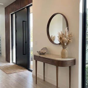 Puede incluir: Una mesa consola de madera con una superficie de piedra clara, con un espejo redondo de marco de madera montado encima. Sobre la mesa hay un jarrón beige con hierba de la pampa seca y un cuenco decorativo plateado. El texto V.O. ARTIS es visible.