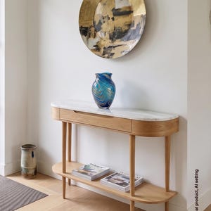 Puede incluir: Una mesa consola de madera clara con tapa de mármol blanco, un cajón y un estante inferior. Un jarrón de cristal azul y dorado se asienta sobre la mesa. Sobre la mesa cuelga un gran plato decorativo de pared redondo y dorado con patrones abstractos.