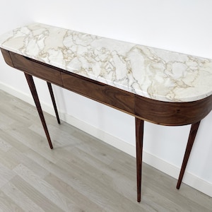 Puede incluir: Una consola de estilo moderno de mediados de siglo con una tapa de mármol y una base de madera oscura. La mesa tiene dos cajones y patas anguladas. La tapa de mármol tiene un patrón de vetas blancas y doradas. La mesa mide unos 152 cm de largo.