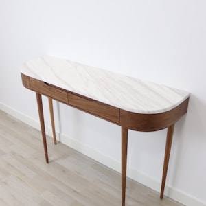 May include: A curved walnut wood console table with two drawers and a white marble top. The table has four slender legs and is shown on a light wood floor.