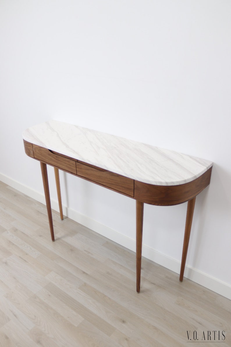 May include: A curved walnut wood console table with two drawers and a white marble top. The table has four slender legs and is shown on a light wood floor.