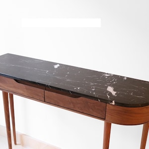 May include: A curved walnut wood console table with two drawers and black marble top. The table has four slender legs.