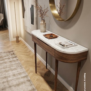 Peut inclure: Une console avec un plateau en marbre blanc et une base en bois incurvée dotée d'un tiroir. La table porte un vase beige avec des branches séchées, un plateau en cuir marron avec des clés et un livre intitulé DESIGN. Le texte V.O. ARTIS est visible.