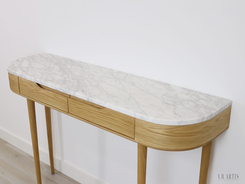 May include: A curved console table with two drawers and a white marble top. The table has four slender legs and is made of light wood.