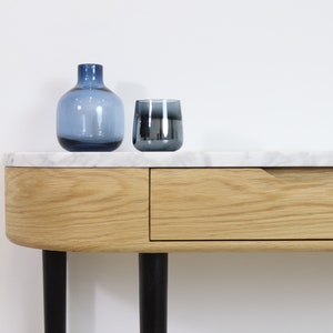 May include: A white marble top console table with a single drawer and black metal legs. The table has a curved front edge and is decorated with a blue glass vase and a blue glass tumbler.