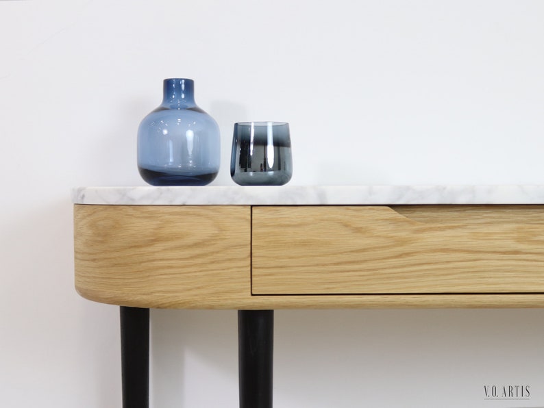 May include: A white marble top console table with a single drawer and black metal legs. The table has a curved front edge and is decorated with a blue glass vase and a blue glass tumbler.