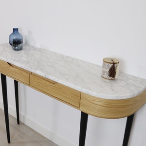 May include: A white marble top console table with two drawers and black metal legs. The table has a curved front edge and is decorated with a blue glass vase and a small decorative box with a tassel.