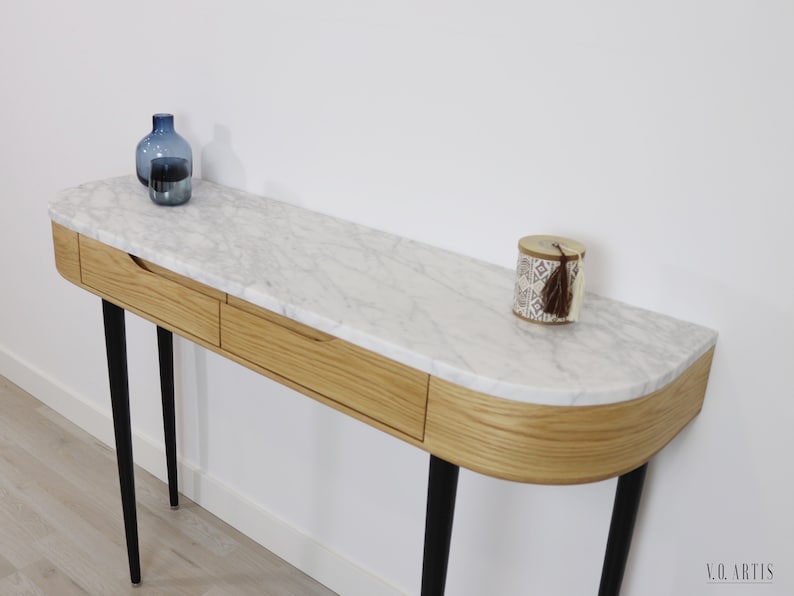 May include: A white marble top console table with two drawers and black metal legs. The table has a curved front edge and is decorated with a blue glass vase and a small decorative box with a tassel.
