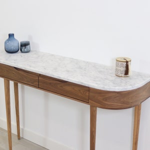 May include: A modern console table with a white marble top and two drawers. The table has a curved front edge and is supported by four slender wooden legs.