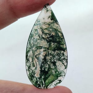 May include: A teardrop-shaped green and white moss agate cabochon. The stone has a swirling pattern of green and white.