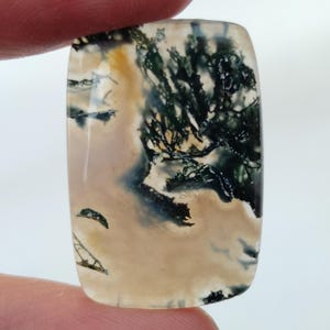 May include: A light brown, green, and black agate cabochon with a dendritic pattern. The stone is cut in a rectangular shape and has a smooth, polished surface.