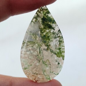 May include: A teardrop-shaped green moss agate cabochon with a white and green pattern. The stone is translucent and has a smooth, polished surface.