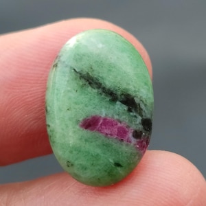 Könnte beinhalten: Ovaler grüner Edelstein mit schwarzen und rosafarbenen Einschlüssen. Der Edelstein ist geschliffen und poliert.