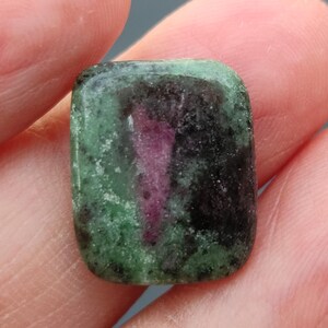 Könnte beinhalten: Ein rechteckiger grüner, schwarzer und rosafarbener Edelstein-Cabochon. Der Stein hat eine raue, strukturierte Oberfläche.