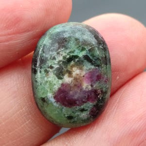 Op de afbeelding: Ovaalvormige groene edelsteen met roze en witte insluitsels. De steen heeft een glad, gepolijst oppervlak.