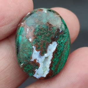 Puede incluir: Gema de forma ovalada con una mezcla de colores verdes, marrones y blancos. La piedra tiene una superficie pulida y un patrón único. La gema se sostiene entre dos dedos.