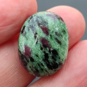 Könnte beinhalten: Ovaler Edelstein mit grüner Grundfarbe, gesprenkelt mit dunkelroten und schwarzen Einschlüssen. Der polierte Stein wird zwischen Fingern gehalten, wodurch seine natürlichen Muster und Texturen hervorgehoben werden. Eine Nahaufnahme zeigt die einzigartigen Farbvariationen.