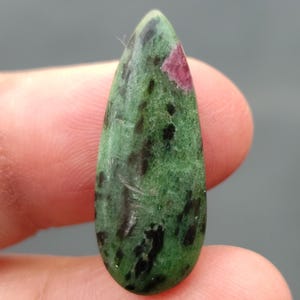 Könnte beinhalten: Ein tropfenförmiger grüner Edelstein mit schwarzen und rosafarbenen Einschlüssen. Der Edelstein ist im Cabochon-Schliff gefasst.