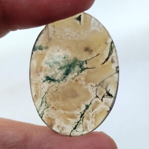 May include: A light brown oval cabochon with green and brown dendritic patterns. The stone is translucent and has a smooth, polished surface.