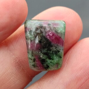 Könnte beinhalten: Ein grüner, rosa und schwarzer Edelstein-Cabochon mit facettierter Form. Der Edelstein ist in einer rechteckigen Form mit abgerundeten Ecken geschnitten.