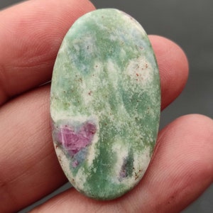Könnte beinhalten: Ovaler grüner und weißer Edelstein-Cabochon mit einer rosa und blauen Einlagerung. Der Stein hat eine gesprenkelte Optik.