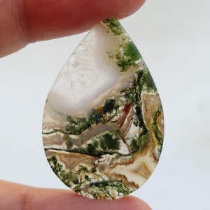 Pode incluir: Um cabochão de ágata verde musgo em forma de lágrima com inclusões brancas, marrons e vermelhas. A pedra tem um padrão natural e orgânico.