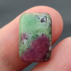 Könnte beinhalten: Ein rechteckiger Edelstein mit abgerundeten Kanten, der eine Mischung aus Grün, tiefem Rot und Weiß aufweist. Die Oberfläche des Steins hat ein gesprenkeltes Aussehen, mit einer Mischung aus Farben und Texturen. Der Edelstein wird zwischen zwei Fingern gehalten.