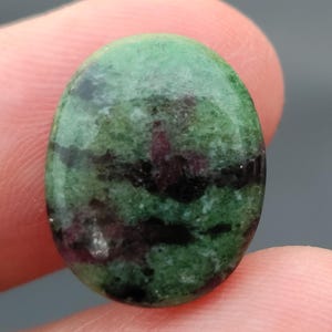 Könnte beinhalten: Ein ovaler Edelstein in Grün-, Schwarz- und Lilatönen mit einer glatten, polierten Oberfläche.