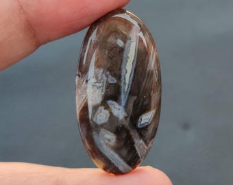 May include: A brown and white agate cabochon with a polished oval shape. The stone has a unique pattern of swirls and lines.