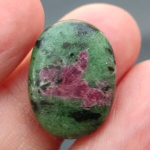 Könnte beinhalten: Ovaler grüner Edelstein mit einer rosa und schwarzen Einlagerung. Der Edelstein ist poliert und hat eine glatte Oberfläche.