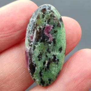 Op de afbeelding: Een ovaalvormige edelsteen met een mix van kleuren. De steen heeft een groene basis met zwarte spikkels en vlekken van roze en wit. De edelsteen wordt tussen vingers gehouden.