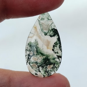 May include: A teardrop-shaped moss agate gemstone. The translucent stone features a mix of white, beige, and green inclusions, creating a natural, organic pattern. The stone is held between two fingers, showcasing its polished surface.