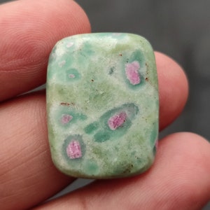 Könnte beinhalten: Ein grüner und rosa Cabochon-Edelstein mit einem gesprenkelten Muster. Der Edelstein ist in rechteckiger Form geschliffen und hat eine glatte, polierte Oberfläche.