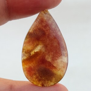 May include: A teardrop-shaped cabochon gemstone with a red, orange, and yellow colour pattern. The stone is translucent and has a cloudy appearance.