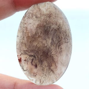 Pode incluir: Um close-up de um cabochão de ágata de forma ovalada de cor cinza claro com um padrão dendrítico de inclusões marrom escuras e pretas. A ágata tem uma aparência translúcida, permitindo que a luz passe por ela.