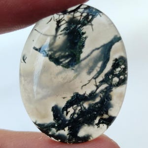 May include: A white agate cabochon with dendritic inclusions in shades of black and green. The stone has a smooth, polished surface and an oval shape.