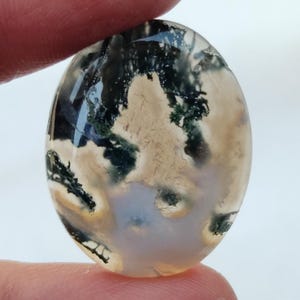 May include: A close-up of a light brown, oval-shaped agate cabochon with green moss inclusions. The agate has a translucent appearance and shows a variety of patterns and textures.