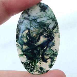 May include: Oval-shaped moss agate gemstone with a translucent white base and intricate green and dark blue patterns. The polished stone is held between fingers, showcasing its natural, organic design. This gemstone is ideal for jewelry making.