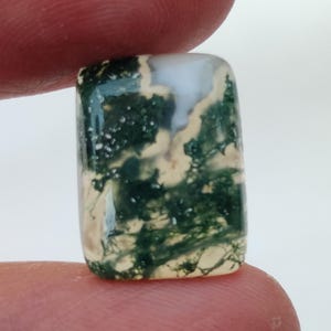 May include: A green moss agate cabochon with a white and green pattern. The stone is rectangular and has a smooth, polished surface.