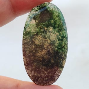 Pode incluir: Cabochão de ágata ovalado de cor verde, marrom e branco com um padrão de redemoinho.