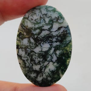 Op de afbeelding: Ovaalvormige groene, witte en zwarte mosagaat cabochon. De steen heeft een draaiende patroon van groen, wit en zwart.