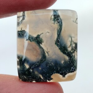 May include: A light gray agate cabochon with a unique dendritic pattern of dark green and black inclusions. The stone has a smooth, polished surface and is rectangular in shape.