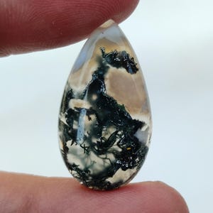 May include: A teardrop-shaped cabochon of moss agate with a white, gray, and green pattern. The stone is translucent and has a smooth, polished surface.