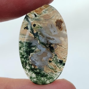 May include: Oval-shaped moss agate gemstone. The stone features a mix of colors, including green, brown, and white, with intricate patterns resembling moss and landscapes. The gemstone is held between two fingers.
